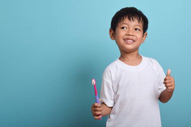Elinde diş fırçası olan mutlu bir Asyalı çocuk mavi arka planda izole bir şekilde baş parmak kaldırıyor. Çocuk dişlerini göstererek gülümsüyor. Çocuk diş bakımı ve hijyen