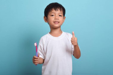 Elinde diş fırçası olan mutlu bir Asyalı çocuk mavi arka planda izole bir şekilde baş parmak kaldırıyor. Çocuk dişlerini göstererek gülümsüyor. Çocuk diş bakımı ve hijyen
