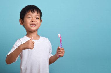 Elinde diş fırçası olan mutlu bir Asyalı çocuk mavi arka planda izole bir şekilde baş parmak kaldırıyor. Çocuk dişlerini göstererek gülümsüyor. Çocuk diş bakımı ve hijyen