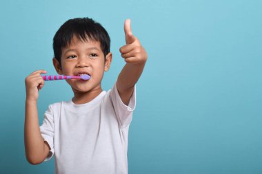 Temiz dişleri olan mutlu bir Asyalı çocuk kamerayı işaret ederken dişlerini fırçalıyor. Mavi arka planda izole edilmiş. Sağlık, ağız temizliği.