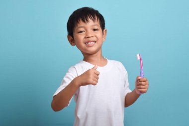 Elinde diş fırçası olan mutlu bir Asyalı çocuk mavi arka planda izole bir şekilde baş parmak kaldırıyor. Çocuk dişlerini göstererek gülümsüyor. Çocuk diş bakımı ve hijyen