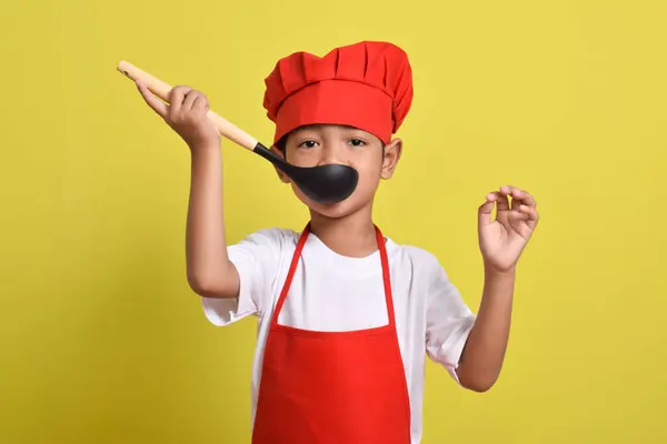 Sarı arka planda kepçe tutan sevimli bir aşçı. Asyalı çocuk yemek yapıyor