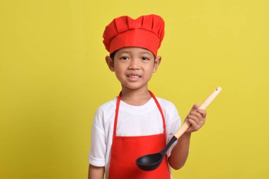 Sarı arka planda kepçe tutan sevimli bir aşçı. Asyalı çocuk yemek yapıyor
