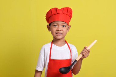 Sarı arka planda kepçe tutan sevimli bir aşçı. Asyalı çocuk yemek yapıyor