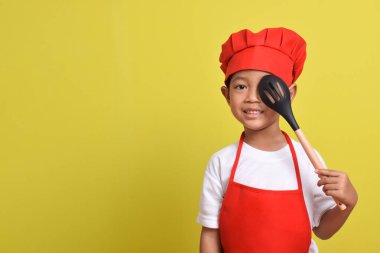 Sevimli küçük şef kepçeyle gözlerini kapatıyor. Sarı arka planda kırmızı önlük ve şapka takan Asyalı çocuk.