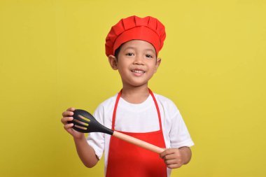 Kaşığı olan mutlu bir aşçı. Sarı arka planda kırmızı önlük ve şapka giyen Asyalı çocuk.