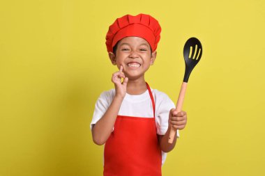Kaşığı olan mutlu bir aşçı. Sarı arka planda kırmızı önlük ve şapka giyen Asyalı çocuk.