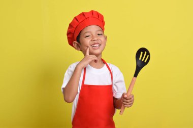 Kaşığı olan mutlu bir aşçı. Sarı arka planda kırmızı önlük ve şapka giyen Asyalı çocuk.