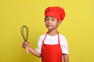 Mutfak çırpıcısını tutan sevimli bir aşçı. Sarı arka planda kırmızı önlük ve şapka giyen Asyalı çocuk.