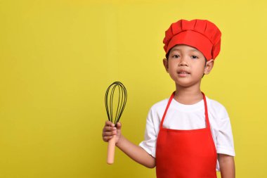 Mutfak çırpıcısını tutan sevimli bir aşçı. Sarı arka planda kırmızı önlük ve şapka giyen Asyalı çocuk.