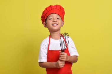 Mutfak çırpıcısını tutan sevimli bir aşçı. Sarı arka planda kırmızı önlük ve şapka giyen Asyalı çocuk.