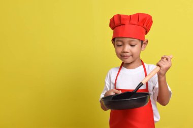Kızartma tavası ve spatulası olan sevimli bir aşçı. Sarı arka planda kırmızı önlük ve şapka giyen Asyalı çocuk.