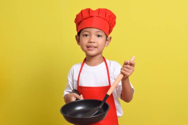 Kızartma tavası ve spatula tutan sevimli bir aşçı. Sarı arka planda kırmızı önlük ve şapka giyen Asyalı çocuk.