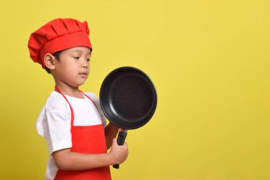 Kızartma tavası ve spatulası olan sevimli bir aşçı. Sarı arka planda kırmızı önlük ve şapka giyen Asyalı çocuk.