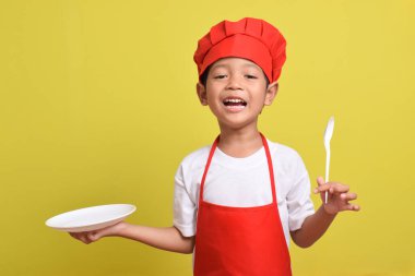 Mutfak tabağı ve kaşığı olan sevimli bir aşçı. Sarı arka planda kırmızı önlük ve şapka giyen Asyalı çocuk.