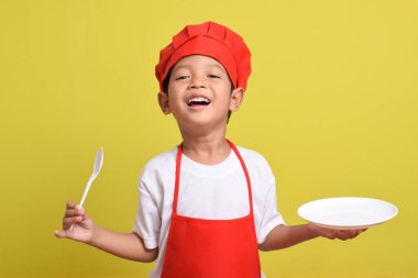 Mutfak tabağı ve kaşığı olan sevimli bir aşçı. Sarı arka planda kırmızı önlük ve şapka giyen Asyalı çocuk.