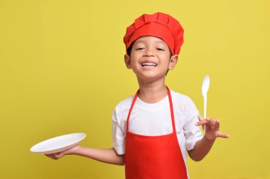 Mutfak tabağı ve kaşığı olan sevimli bir aşçı. Sarı arka planda kırmızı önlük ve şapka giyen Asyalı çocuk.