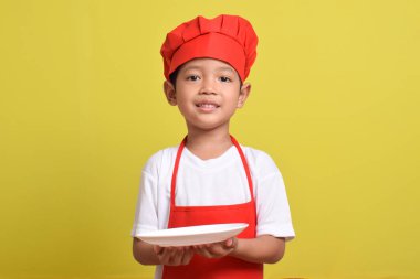 Tabağı olan sevimli bir aşçı. Sarı arka planda kırmızı önlük ve şapka takan Asyalı çocuk. Yemek servisi kavramı.