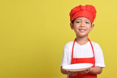 Tabağı olan sevimli bir aşçı. Sarı arka planda kırmızı önlük ve şapka takan Asyalı çocuk. Yemek servisi kavramı.