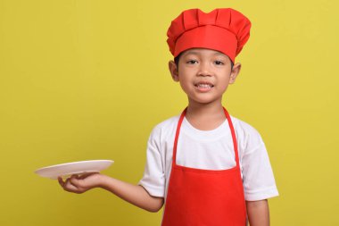 Tabağı olan sevimli bir aşçı. Sarı arka planda kırmızı önlük ve şapka takan Asyalı çocuk. Yemek servisi kavramı.