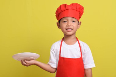 Tabağı olan sevimli bir aşçı. Sarı arka planda kırmızı önlük ve şapka takan Asyalı çocuk. Yemek servisi kavramı.
