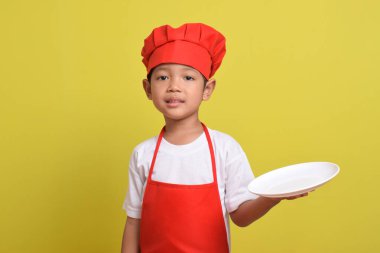 Tabağı olan sevimli bir aşçı. Sarı arka planda kırmızı önlük ve şapka takan Asyalı çocuk. Yemek servisi kavramı.