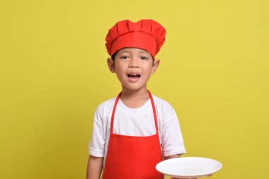 Tabağı olan sevimli bir aşçı. Sarı arka planda kırmızı önlük ve şapka takan Asyalı çocuk. Yemek servisi kavramı.
