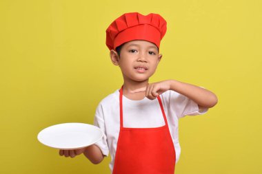 Parmakla tabağı gösteren sevimli bir aşçı. Sarı arka planda kırmızı önlük ve şapka takan Asyalı çocuk. Yemek servisi kavramı.