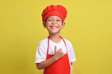 Sarı arka planda baş parmak gösteren sevimli bir aşçı. Asyalı çocuk kırmızı önlük ve şapka takıyor. Harika küçük şef.