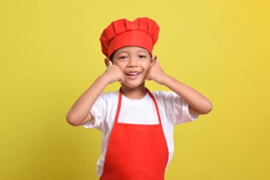 Sarı arka planda baş parmak gösteren sevimli bir aşçı. Asyalı çocuk kırmızı önlük ve şapka takıyor. Harika küçük şef.