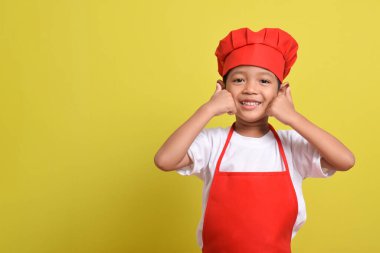Sarı arka planda baş parmak gösteren sevimli bir aşçı. Asyalı çocuk kırmızı önlük ve şapka takıyor. Harika küçük şef.