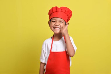 Sarı arka planda baş parmak gösteren sevimli bir aşçı. Asyalı çocuk kırmızı önlük ve şapka takıyor. Harika küçük şef.