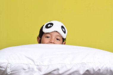 Sarı arka planda, yastığa bırakılmış pijama ve uyku maskesi takan sevimli Asyalı çocuğun portresi. Sağlıklı uyku konsepti. Boşluğu kopyala