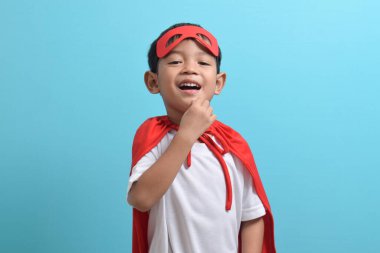 Süper kahraman kostümlü Asyalı çocuğun portresi. Mavi arka planda izole edilmiş kameraya bakıyor.