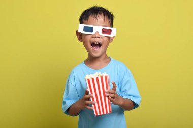 Heyecanlı Asyalı gözlüklü çocuk sarı arka planda izole edilmiş patlamış mısır kovası tutarken film izliyor. Film ve sinema izleme kavramı