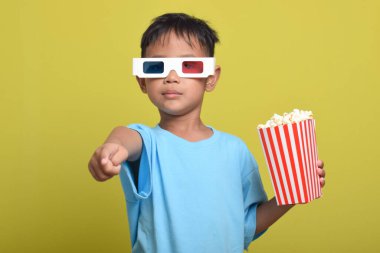 Üç boyutlu gözlük takan Asyalı çocuk sarı arka planda izole edilmiş patlamış mısır kovası tutarken film izliyor. Kamerayı işaret eden parmak. Film ve sinema izleme kavramı