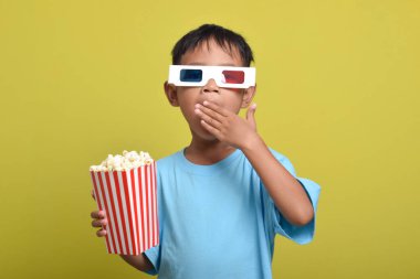 Asyalı çocuk şokta ve elleri ağzı kapatıyor, 3D gözlük takıyor, sarı arka planda izole edilmiş patlamış mısır kovası tutarken film izliyor. Kamerayı işaret eden parmak. Film ve sinema konsepti