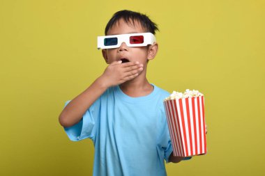 Asyalı çocuk şokta ve elleri ağzı kapatıyor, 3D gözlük takıyor, sarı arka planda izole edilmiş patlamış mısır kovası tutarken film izliyor. Kamerayı işaret eden parmak. Film ve sinema konsepti