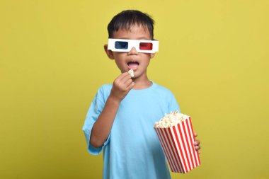 Asyalı çocuk 3D gözlük takıyor, patlamış mısır yiyor ve sarı arka planda izole edilmiş film izliyor. Film ve sinema konsepti izlemek