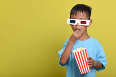 Asyalı çocuk 3D gözlük takıyor, patlamış mısır yiyor ve sarı arka planda izole edilmiş film izliyor. Film ve sinema konsepti izlemek