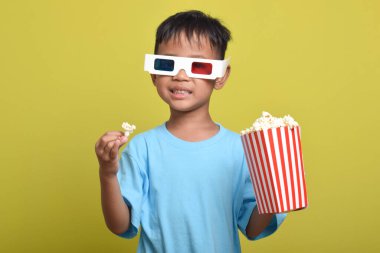 Asyalı çocuk 3D gözlük takıyor, patlamış mısır yiyor ve sarı arka planda izole edilmiş film izliyor. Film ve sinema konsepti izlemek