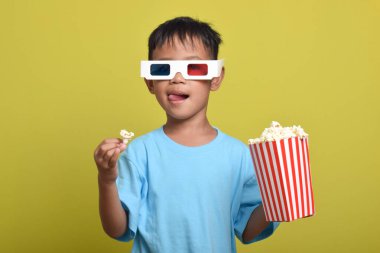 Asyalı çocuk 3D gözlük takıyor, patlamış mısır yiyor ve sarı arka planda izole edilmiş film izliyor. Film ve sinema konsepti izlemek