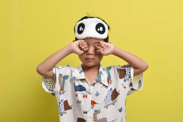 Uykulu Asyalı çocuğun portresi sarı arka planda gözlerini ovuşturuyor. Pijama ve uyku maskesi takan bir çocuk. Sağlıklı uyku konsepti. Boşluğu kopyala
