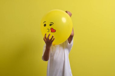 Öpücük emojisi. Yakışıklı Asyalı çocuk sarı arka planda izole edilmiş öpücük simgesi balonunun arkasına saklanıyor.
