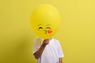 Öpücük emojisi. Yakışıklı Asyalı çocuk sarı arka planda izole edilmiş romantik duygu balonlarının arkasına saklanıyor.