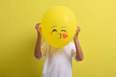 Öpücük emojisi. Yakışıklı Asyalı çocuk sarı arka planda izole edilmiş romantik duygu balonlarının arkasına saklanıyor.