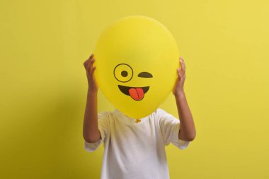 Dil emojisi. Sarı arka planda izole edilmiş dilini dışarı çıkaran balon simgesinin arkasına saklanan sevimli Asyalı çocuk. Kapalı gözlerle alay eden emoji