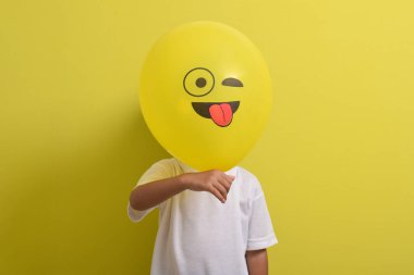 Dil emojisi. Sarı arka planda izole edilmiş dilini dışarı çıkaran balon simgesinin arkasına saklanan sevimli Asyalı çocuk. Kapalı gözlerle alay eden emoji