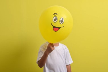 Gülümse emoji. Yakışıklı Asyalı çocuk sarı arka planda izole edilmiş mutlu duygu balonunun arkasına saklanıyor.