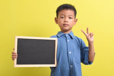 Sarı arka planda izole edilmiş boş bir tebeşir tahtası tutan sevimli küçük Asyalı çocuk. Metin, kopyalama alanı, biçimlendirme pankartı için boşluk